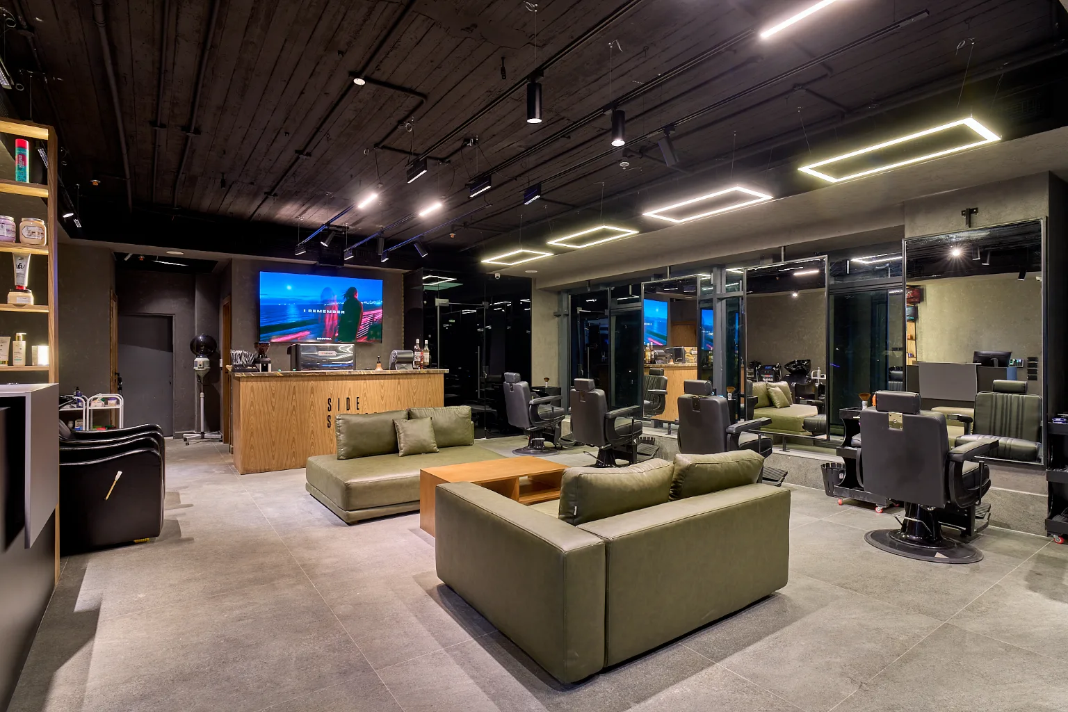 KK Barbershop, Sheikh Zayed, as built: olive-green leather sofas around a wooden coffee table, barber chairs facing mirrored walls, and a dark timber-board ceiling with square and linear lights.