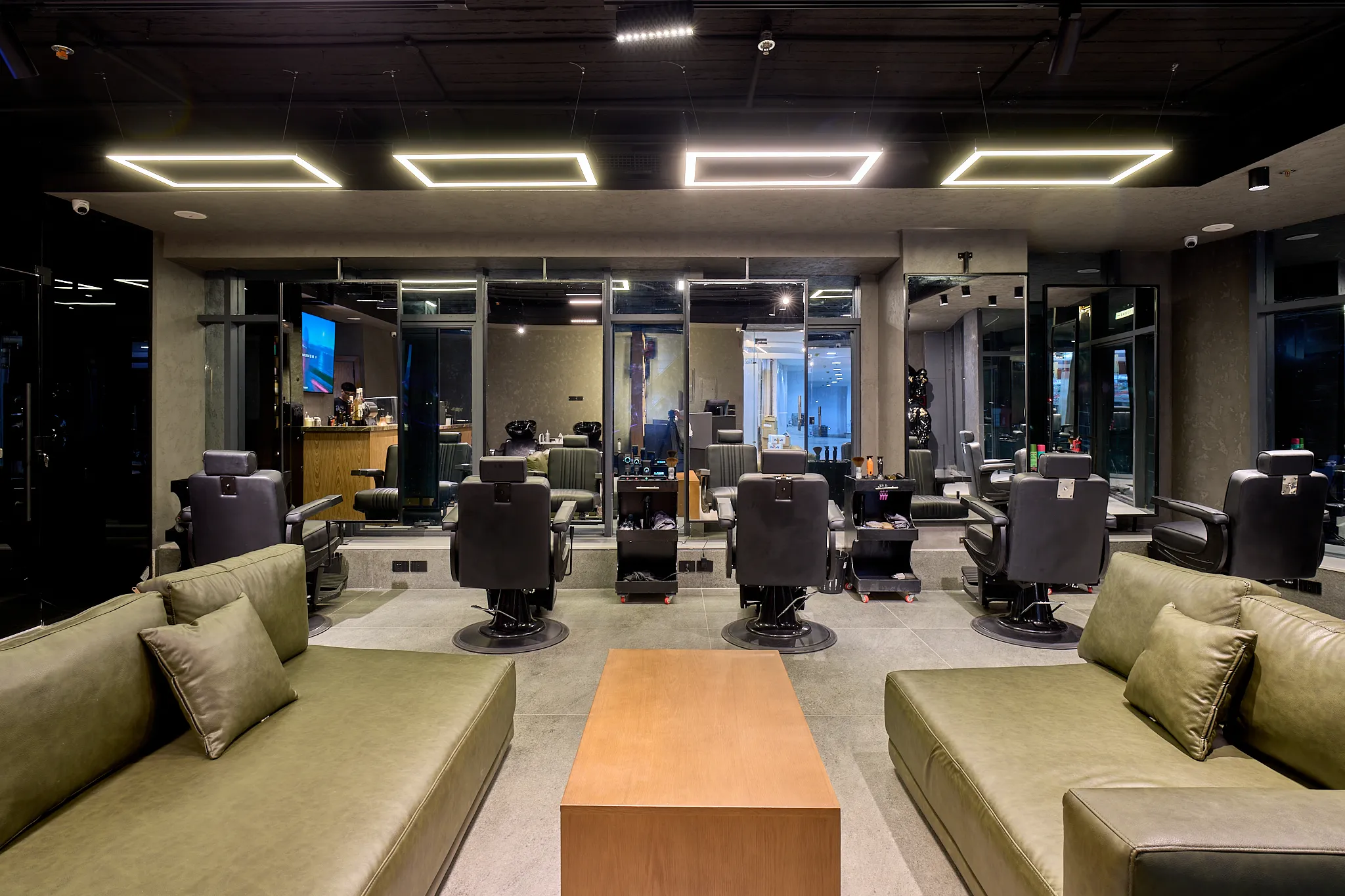 Front view of the barber stations: black barber chairs and trolleys facing the mirrors, seen between two olive-green sofas and a long wooden coffee table.