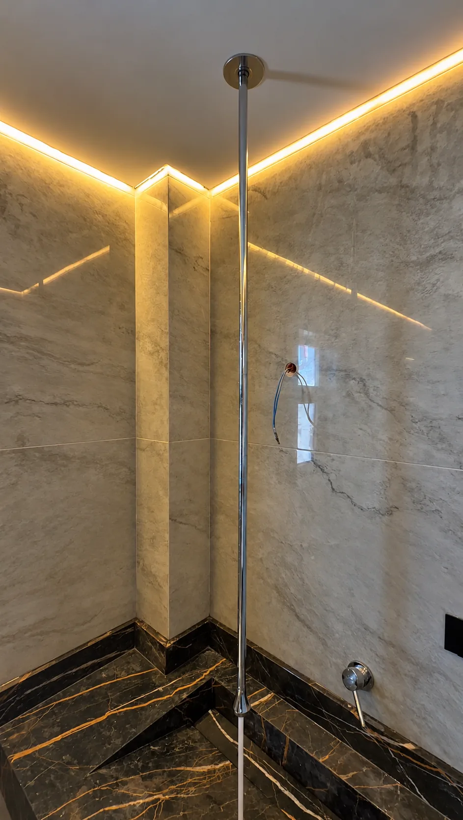A shower area in light veined stone with a dark veined floor and a lit cove, with its fittings still exposed.