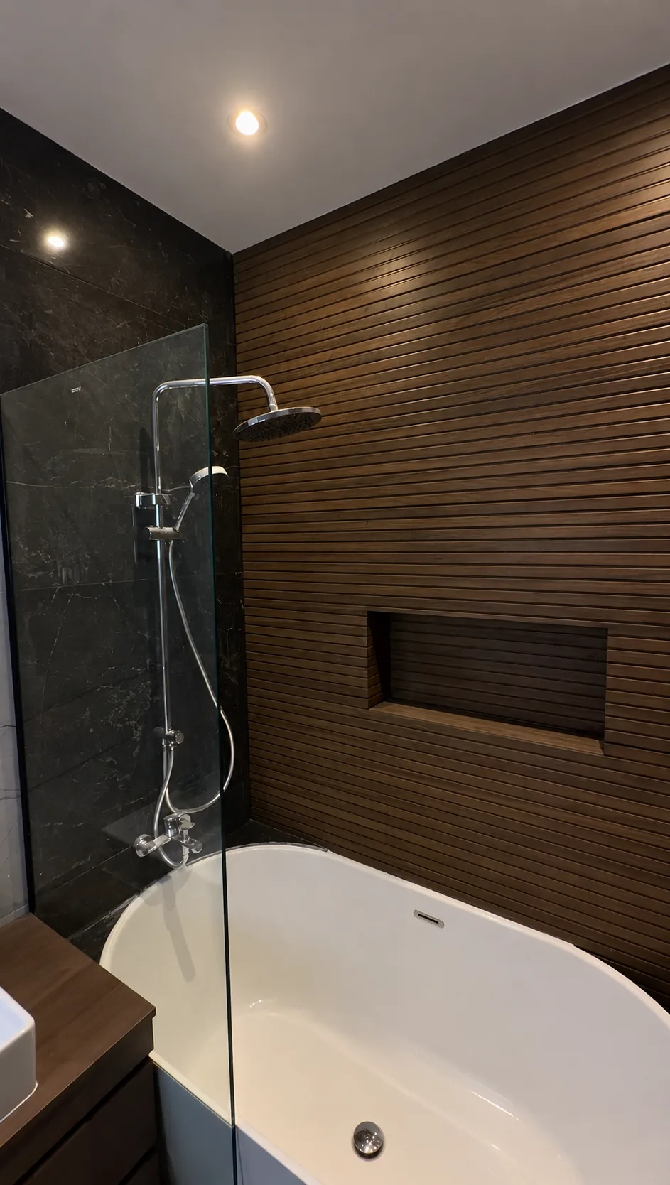 A bathroom with a freestanding bathtub, a glass shower screen and a dark wood-slat wall.